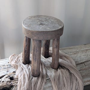 Könnte beinhalten: Ein kleiner, rustikaler Holzschemel mit rundem Sitz und drei stabilen Beinen. Der Schemel ist aus verwittertem Holz gefertigt und zeigt natürliche Maserung und Textur. Er wird auf einem Treibholzstück mit einem fließenden, hellfarbigen Stoff drapiert präsentiert.