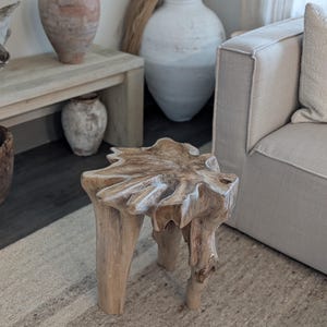 May include: A natural wood side table with a unique, organic shape. The table has three sturdy legs and a textured top. The light brown color complements the neutral tones of the room, creating a rustic and earthy aesthetic. The table is a decorative accent piece.