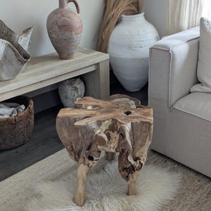 May include: A unique, natural wood side table with a raw, organic design. The table has a rough-hewn top and three sturdy legs, showcasing the natural grain and texture of the wood. It sits on a fluffy, white rug, adding a touch of softness to the space.
