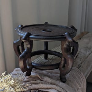 May include: Dark brown wooden display stand with a circular top and ornate, curved legs. The stand has a dark finish and is designed to hold a decorative object. The stand is approximately 6 inches in diameter.