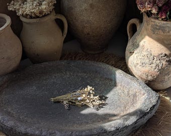 May include: A large, dark grey stone bowl with a small bunch of dried flowers. Several antique, weathered clay vases of varying sizes are in the background, some holding dried flowers. The scene is set on a woven mat, creating a natural, earthy aesthetic.