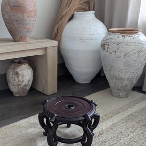 Puede incluir: Un soporte de madera marr&oacute;n oscuro y ornamentado con una parte superior circular y patas curvas. Varias jarrones de cer&aacute;mica grandes y texturizados en tonos de blanco, beige y marr&oacute;n se encuentran en el fondo. La escena est&aacute; ambientada sobre una alfombra de tonos neutros.
