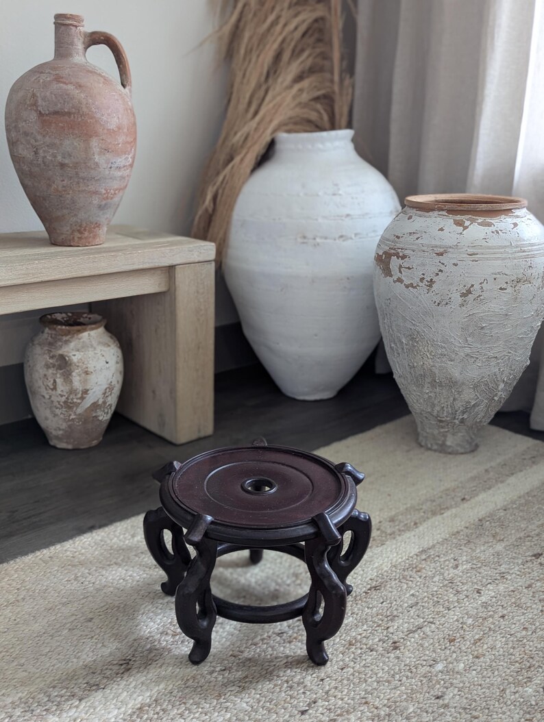 Puede incluir: Un soporte de madera marr&oacute;n oscuro y ornamentado con una parte superior circular y patas curvas. Varias jarrones de cer&aacute;mica grandes y texturizados en tonos de blanco, beige y marr&oacute;n se encuentran en el fondo. La escena est&aacute; ambientada sobre una alfombra de tonos neutros.