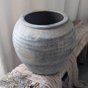 May include: A large, round, gray-toned ceramic pot with a wide opening. The pot has a textured, weathered appearance with horizontal banding. It sits on a rustic wooden surface with a draped fabric and dried flowers.