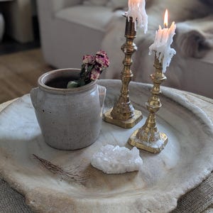 May include: A decorative arrangement featuring a stone bowl, a small ceramic pot with dried flowers, a white crystal, and two lit brass candlesticks with dripping wax. The items are arranged on a neutral-toned surface.