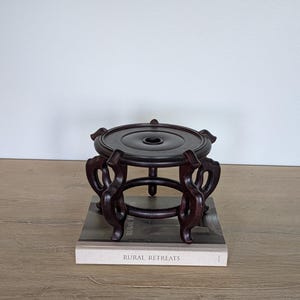 Puede incluir: Un soporte de madera marr&oacute;n oscuro y ornamentado con una parte superior circular y patas curvas. El soporte est&aacute; sobre un libro titulado "RURAL RETREATS". El soporte est&aacute; dise&ntilde;ado para sostener un objeto decorativo.