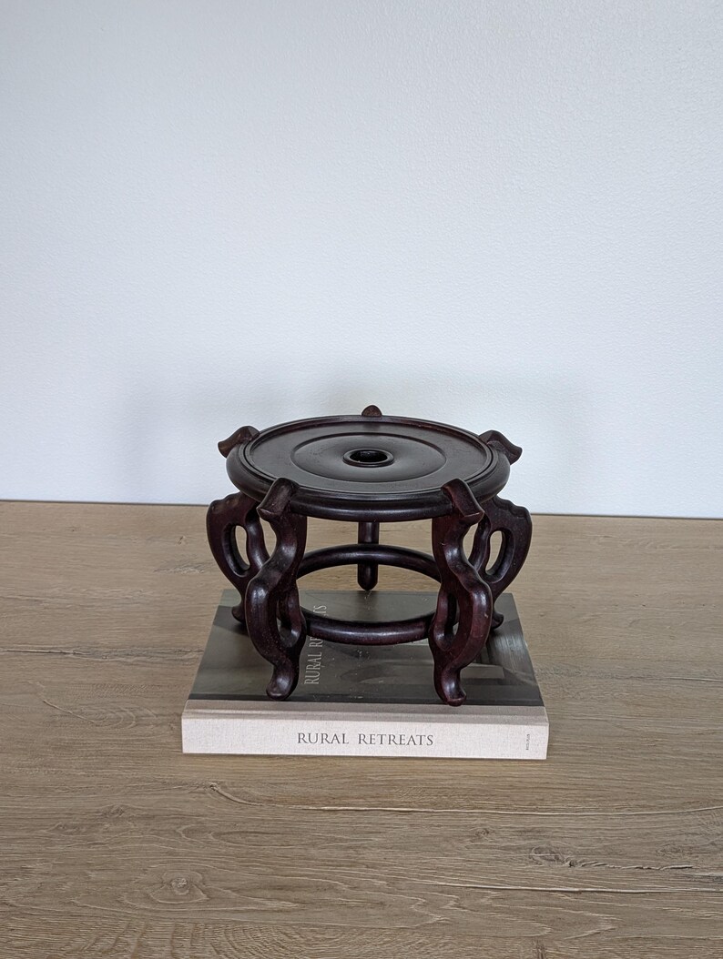 Puede incluir: Un soporte de madera marr&oacute;n oscuro y ornamentado con una parte superior circular y patas curvas. El soporte est&aacute; sobre un libro titulado "RURAL RETREATS". El soporte est&aacute; dise&ntilde;ado para sostener un objeto decorativo.