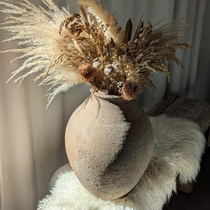 May include: A large, textured, earthenware vase filled with dried flowers and grasses. The arrangement includes feathery pampas grass, seed pods, and other dried elements in shades of beige and brown. The vase sits on a wooden bench covered with a white, fluffy sheepskin.