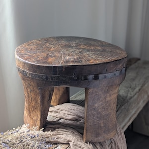 May include: A rustic, round wooden stool with a dark brown finish. The stool has a circular top and three sturdy legs. A metal band with rivets encircles the top. The wood grain is visible. The stool is approximately 12 inches in height.