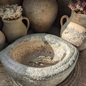 May include: A weathered, grey stone bowl with a rough texture, containing a small bunch of dried flowers. The bowl is surrounded by several antique, earthen-toned vases, some with dried floral arrangements. The scene evokes a rustic, vintage aesthetic.