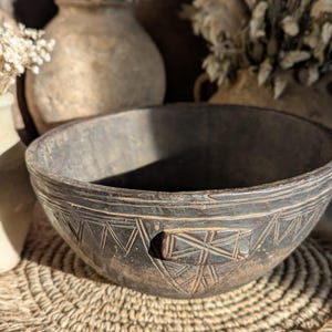 May include: A dark brown, hand-carved wooden bowl with geometric patterns around the rim and a small handle. The bowl sits on a woven mat, with dried flowers and vases in the background.