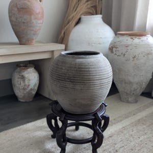 Puede incluir: Un jarr&oacute;n de cer&aacute;mica gris y acanalado se encuentra sobre un soporte de madera oscura con patas ornamentadas. Varios otros jarrones de cer&aacute;mica envejecidos de varios tama&ntilde;os y colores se encuentran en el fondo, creando una exhibici&oacute;n r&uacute;stica de tonos neutros. Las hierbas secas a&ntilde;aden un elemento natural.