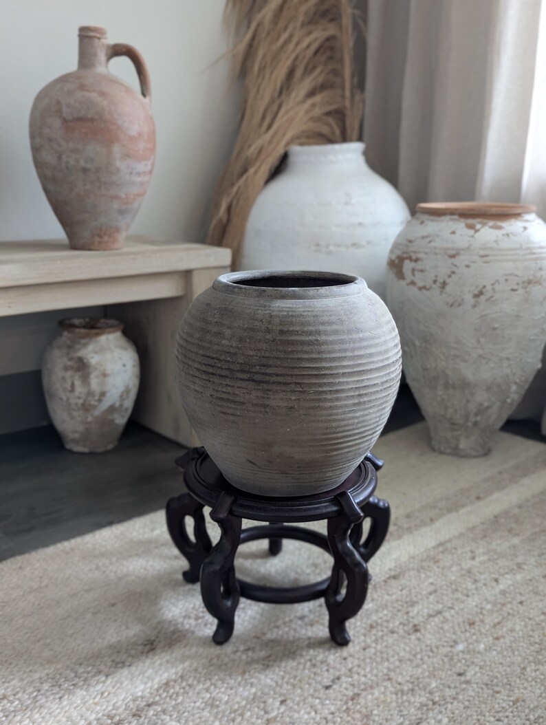 Puede incluir: Un jarr&oacute;n de cer&aacute;mica gris y acanalado se encuentra sobre un soporte de madera oscura con patas ornamentadas. Varios otros jarrones de cer&aacute;mica envejecidos de varios tama&ntilde;os y colores se encuentran en el fondo, creando una exhibici&oacute;n r&uacute;stica de tonos neutros. Las hierbas secas a&ntilde;aden un elemento natural.