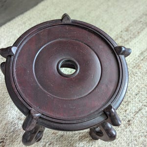 Puede incluir: Un soporte de exhibici&oacute;n de madera marr&oacute;n oscuro con una parte superior circular y tres patas decorativas. El soporte tiene un agujero central y est&aacute; dise&ntilde;ado para sostener un objeto peque&ntilde;o. La madera tiene un acabado rico y pulido.
