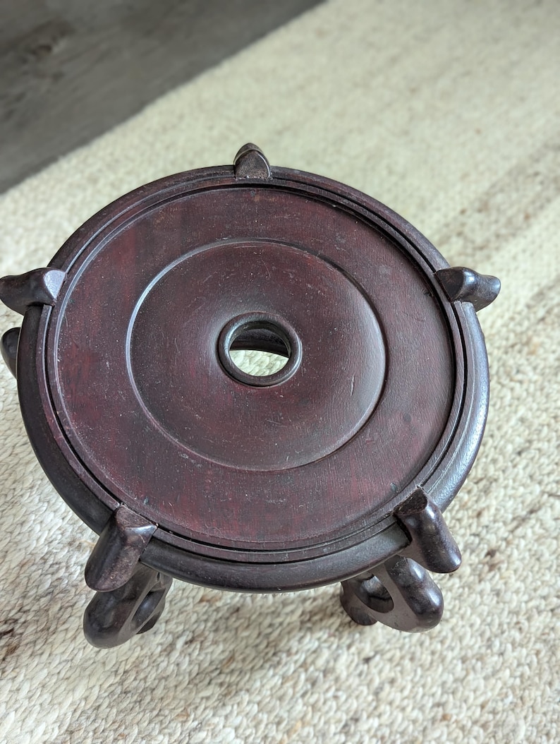 Puede incluir: Un soporte de exhibici&oacute;n de madera marr&oacute;n oscuro con una parte superior circular y tres patas decorativas. El soporte tiene un agujero central y est&aacute; dise&ntilde;ado para sostener un objeto peque&ntilde;o. La madera tiene un acabado rico y pulido.