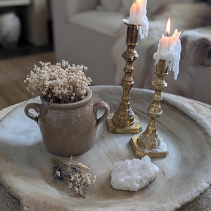 Op de afbeelding: Een decoratieve opstelling in een stenen kom. Het bevat een kleine keramische pot met gedroogde bloemen, een cluster van kwartskristallen en twee messing kandelaars met brandende kaarsen. De kaarsen hebben gesmolten was.