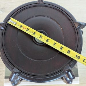 Puede incluir: Soporte de exhibici&oacute;n de madera marr&oacute;n oscuro con una parte superior circular y cuatro patas curvas. Una cinta m&eacute;trica amarilla muestra que el soporte tiene aproximadamente 25 cm de di&aacute;metro. El soporte tiene un agujero central y est&aacute; dise&ntilde;ado para sostener un objeto decorativo.