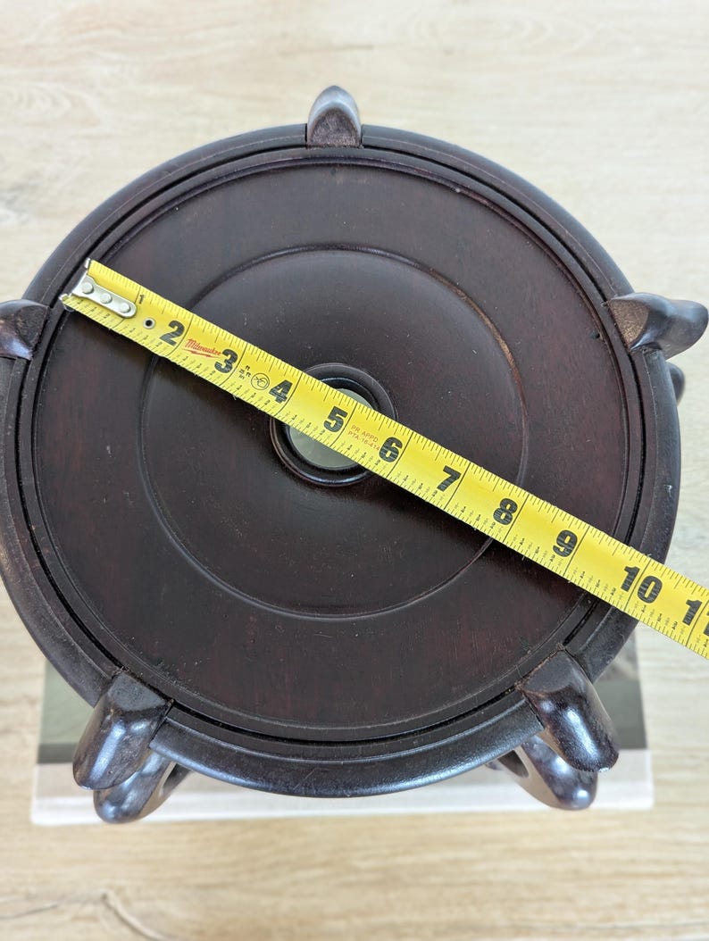 Puede incluir: Soporte de exhibici&oacute;n de madera marr&oacute;n oscuro con una parte superior circular y cuatro patas curvas. Una cinta m&eacute;trica amarilla muestra que el soporte tiene aproximadamente 25 cm de di&aacute;metro. El soporte tiene un agujero central y est&aacute; dise&ntilde;ado para sostener un objeto decorativo.