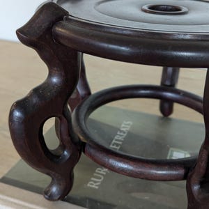 Puede incluir: Un soporte de madera marr&oacute;n oscuro de estilo antiguo, con una parte superior circular y un agujero central. El soporte tiene tres patas curvas y un anillo decorativo en la base. La madera tiene un acabado pulido.
