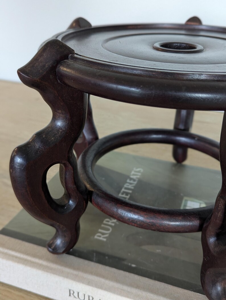 Puede incluir: Un soporte de madera marr&oacute;n oscuro de estilo antiguo, con una parte superior circular y un agujero central. El soporte tiene tres patas curvas y un anillo decorativo en la base. La madera tiene un acabado pulido.