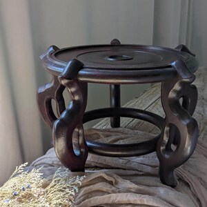 Puede incluir: Un soporte de madera marr&oacute;n oscuro para plantas con una parte superior circular y patas decorativas y curvas. El soporte tiene un agujero central y un anillo inferior para mayor estabilidad. La madera tiene un acabado rico y pulido.