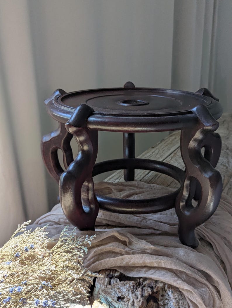 Puede incluir: Un soporte de madera marr&oacute;n oscuro para plantas con una parte superior circular y patas decorativas y curvas. El soporte tiene un agujero central y un anillo inferior para mayor estabilidad. La madera tiene un acabado rico y pulido.