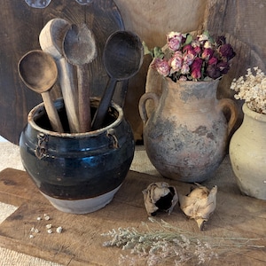 Puede incluir: Naturaleza muerta rústica con utensilios de cocina vintage. Una olla de cerámica azul oscuro y blanca contiene cucharas de madera. Dos jarrones de cerámica envejecidos exhiben flores secas. El arreglo descansa sobre una superficie de madera desgastada.