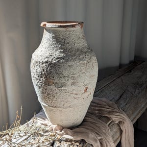 Puede incluir: Un jarrón grande de estilo antiguo con un exterior texturizado blanquecino. El jarrón tiene un cuerpo ancho que se estrecha hacia la base y un borde ligeramente ensanchado. Se encuentra sobre una superficie de madera envejecida, con una tela drapeada.