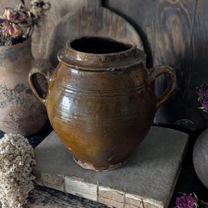 May include: A brown, antique ceramic jar with two handles and a lid. The jar is sitting on a weathered book. The background includes other vintage items, such as a vase with dried flowers and wooden cutting boards.
