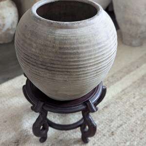 Puede incluir: Una maceta redonda de cer&aacute;mica de color claro con estr&iacute;as horizontales, colocada sobre un soporte de madera marr&oacute;n oscuro y ornamentado. La maceta tiene una abertura ancha y una superficie texturizada, lo que sugiere una est&eacute;tica r&uacute;stica o antigua. El soporte tiene patas curvas.