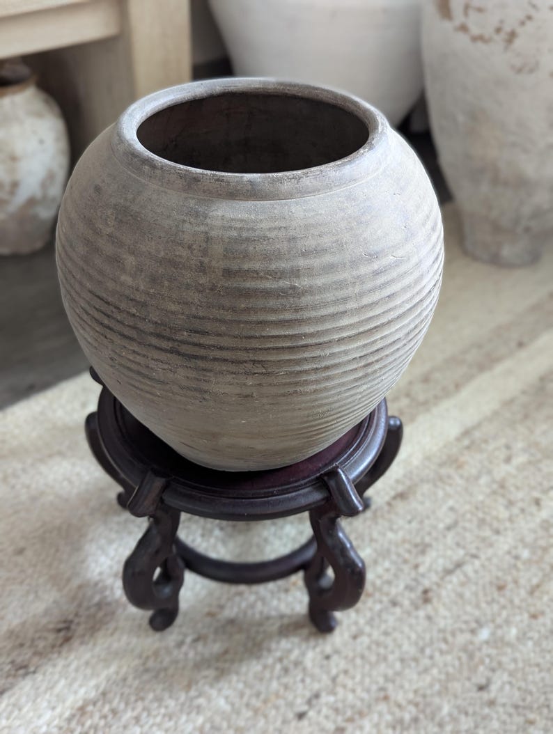 Puede incluir: Una maceta redonda de cer&aacute;mica de color claro con estr&iacute;as horizontales, colocada sobre un soporte de madera marr&oacute;n oscuro y ornamentado. La maceta tiene una abertura ancha y una superficie texturizada, lo que sugiere una est&eacute;tica r&uacute;stica o antigua. El soporte tiene patas curvas.
