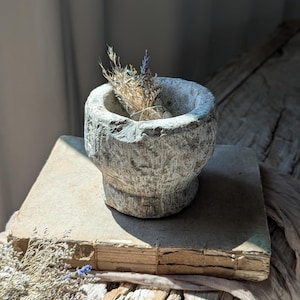 May include: A grey stone mortar, containing dried flowers, sits on an old book. The mortar has a rough, textured surface. The book rests on a wooden surface, with a soft, natural light illuminating the scene.