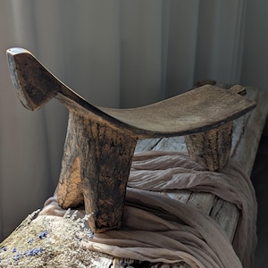 May include: A handcrafted wooden stool with a curved seat and three sturdy legs. The stool has a natural, weathered appearance with visible wood grain and a dark, aged finish. It is placed on a rustic wooden surface with a draped fabric.