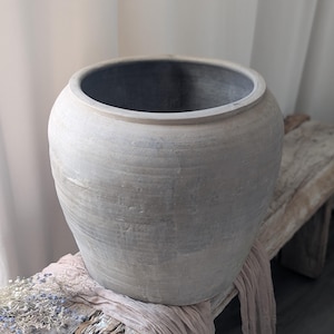 May include: A large, round, gray ceramic pot with a wide opening. The pot has a textured surface and sits on a rustic wooden bench. A light pink fabric and dried flowers are draped around the base of the pot. Home decor.