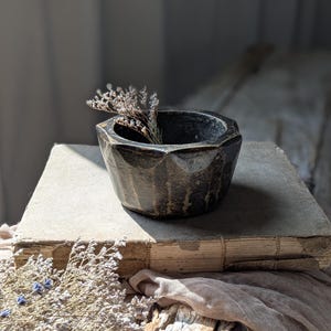 May include: A dark, faceted bowl with a dried floral arrangement inside. The bowl sits atop an aged book with a neutral-colored cloth draped nearby. The scene is lit by natural light, creating shadows and highlighting the textures of the objects.