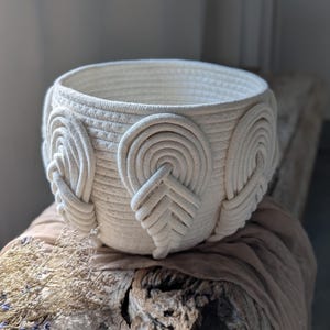 May include: A round, off-white basket constructed from coiled cotton rope. The exterior features a repeating decorative pattern of raised, arch-shaped rope loops arranged in a vertical, leaf-like formation around the circumference of the vessel.