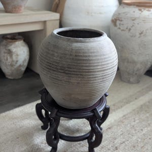 Puede incluir: Un jarr&oacute;n de cer&aacute;mica gris y acanalado con interior oscuro se asienta sobre un soporte de madera oscura y ornamentada. El jarr&oacute;n es redondo con una amplia abertura. Otros jarrones de cer&aacute;mica envejecida est&aacute;n en el fondo.