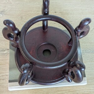 Puede incluir: Un soporte de exhibici&oacute;n de madera marr&oacute;n oscuro con una parte superior e inferior circulares. El soporte tiene un anillo alrededor de la parte superior y cuatro soportes decorativos y curvos. El soporte est&aacute; dise&ntilde;ado para sostener un objeto decorativo.
