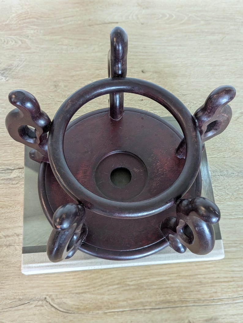 Puede incluir: Un soporte de exhibici&oacute;n de madera marr&oacute;n oscuro con una parte superior e inferior circulares. El soporte tiene un anillo alrededor de la parte superior y cuatro soportes decorativos y curvos. El soporte est&aacute; dise&ntilde;ado para sostener un objeto decorativo.