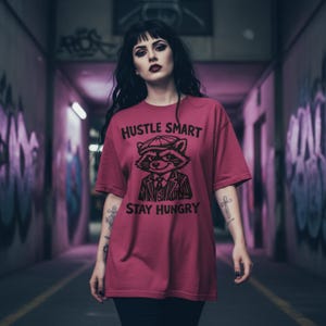 Peut inclure: T-shirt bordeaux avec l'inscription "Hustle Smart Stay Hungry" et un dessin de raton laveur en chapeau et costume. Le t-shirt à manches courtes a une coupe ample.