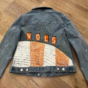May include: A blue denim jacket featuring white paint splatters across the back. The back panel displays the orange text VOLS above a curved graphic design consisting of alternating white and orange vertical stripes with a textured appearance.