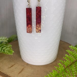 May include: A pair of dangle earrings with red and gold resin bar pendants. The earrings are hanging from silver hooks.
