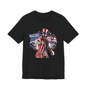 May include: A black t-shirt featuring a graphic of a boxer wearing a red, white, and blue striped top hat and matching boxing trunks. The boxer is throwing a punch, and the text "LIVING IN AMERICA" is displayed in bold blue and red letters.