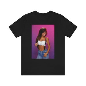 May include: Black t-shirt featuring a framed image of a person wearing a white top, blue jeans, and suspenders. The background is a gradient of pink and purple. The person is smiling and holding the suspenders.