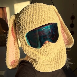 Op de afbeelding: Een crèmekleurige, gehaakte konijnenmuts met lange, slappe oren en roze details aan de binnenkant. De hoed heeft een nekwarmer en wordt gecombineerd met blauw getinte skibrillen. Het woord "goodr" is zichtbaar op de bril.