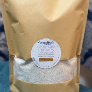 May include: A brown paper bag filled with white goat milk laundry powder. The bag has a white label with the text "Goat Milk Laundry Powder Eucalyptus & Lemon" and "Bright Citrus Scent & Minimal Ingredients".