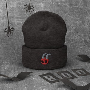 Puede incluir: Gorro negro con una calavera roja y cuernos bordados en la parte delantera. El gorro está sobre una superficie gris con murciélagos de papel negro y la palabra "BOO" en letras negras.