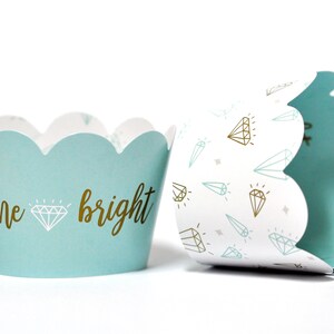 May include: Two light blue cupcake liners with scalloped edges. One liner has a white background with a pattern of gold and light blue diamonds. The other liner has the text "shine bright" in gold lettering with a gold diamond in between the words.