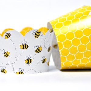 May include: Two cupcake liners with a bee theme. One liner is white with yellow and black bees and dotted flight paths. The other liner is yellow with a honeycomb pattern. The liners have a scalloped edge.