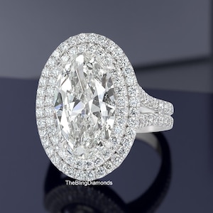Pode incluir: Um anel de noivado deslumbrante com um grande diamante central de corte oval, rodeado por um halo duplo de diamantes menores. O anel tem um design de haste dividida e é cravejado em metal branco. O logotipo The Bling Diamonds é visível.